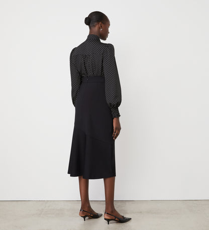 A woman stands facing away against a white wall, wearing a black blouse with white polka dots, the Grace Black Ponte Jersey Midi Skirt with an elasticated waistband, and black flat shoes with a strap.