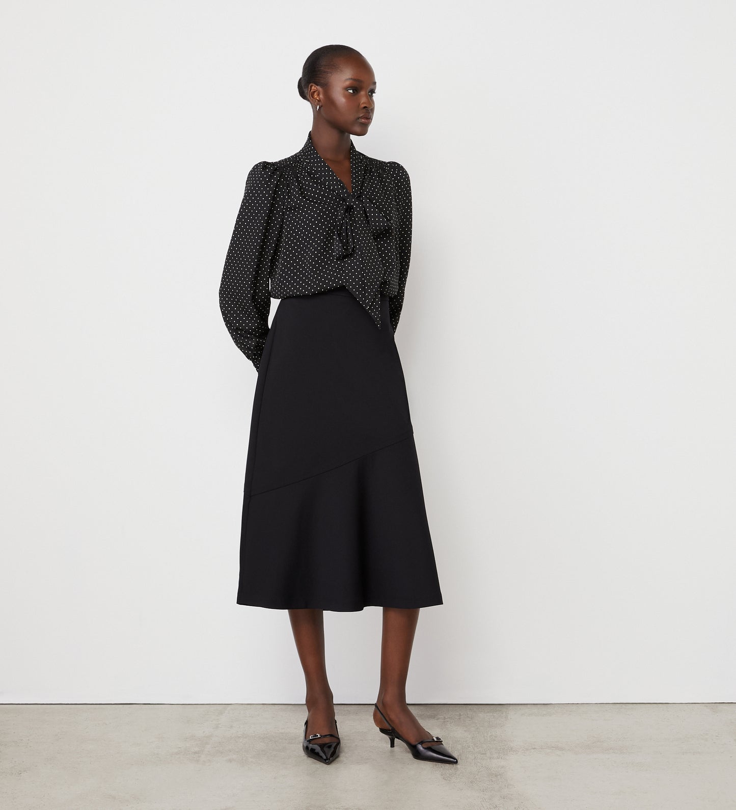 A woman stands against a white wall wearing a black, long-sleeved, polka dot blouse with a bow at the neck and the Grace Black Ponte Jersey Midi Skirt featuring an elasticated waistband. She completes the look with black pointed-toe heels.