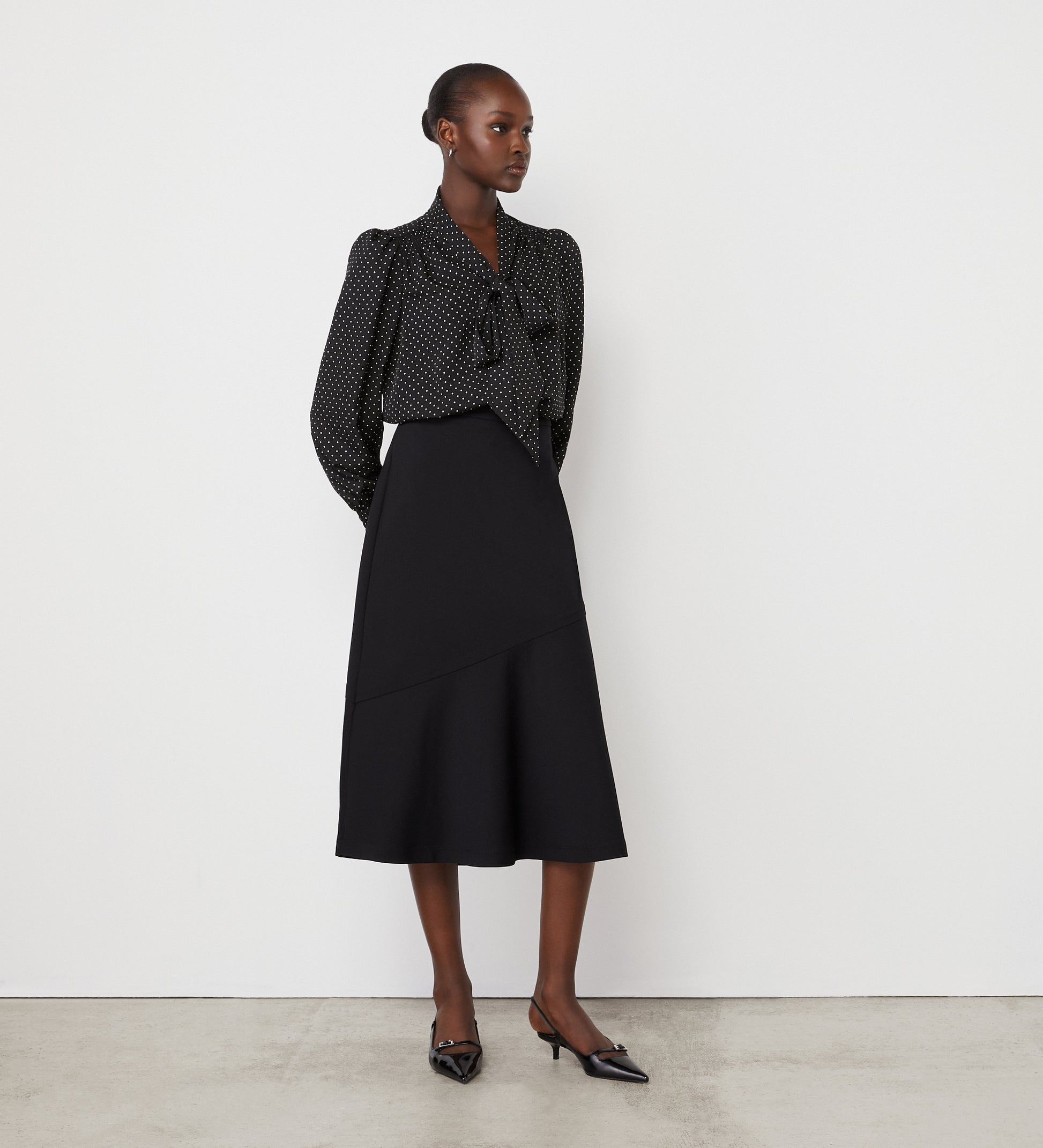 A woman stands against a white wall wearing a black, long-sleeved, polka dot blouse with a bow at the neck and the Grace Black Ponte Jersey Midi Skirt featuring an elasticated waistband. She completes the look with black pointed-toe heels.