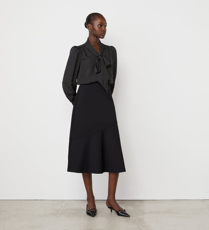 A woman stands against a white wall wearing a black, long-sleeved, polka dot blouse with a bow at the neck and the Grace Black Ponte Jersey Midi Skirt featuring an elasticated waistband. She completes the look with black pointed-toe heels.