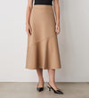 A woman stands against a white wall wearing a black sleeveless top, the Grace Camel Ponte Jersey Midi Skirt with an angled seam, and black pointed-toe heels—timeless pieces ideal for versatile styling.