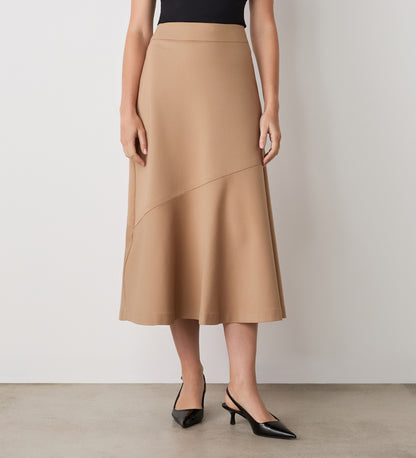 A woman stands against a white wall wearing a black sleeveless top, the Grace Camel Ponte Jersey Midi Skirt with an angled seam, and black pointed-toe heels—timeless pieces ideal for versatile styling.