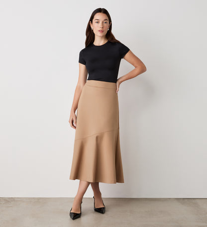 A woman stands against a plain light background, wearing a fitted black short-sleeve top and the Grace Camel Ponte Jersey Midi Skirt with a flared hem. She pairs them with black pointed-toe heels and wears a neutral expression—classic wardrobe essentials.