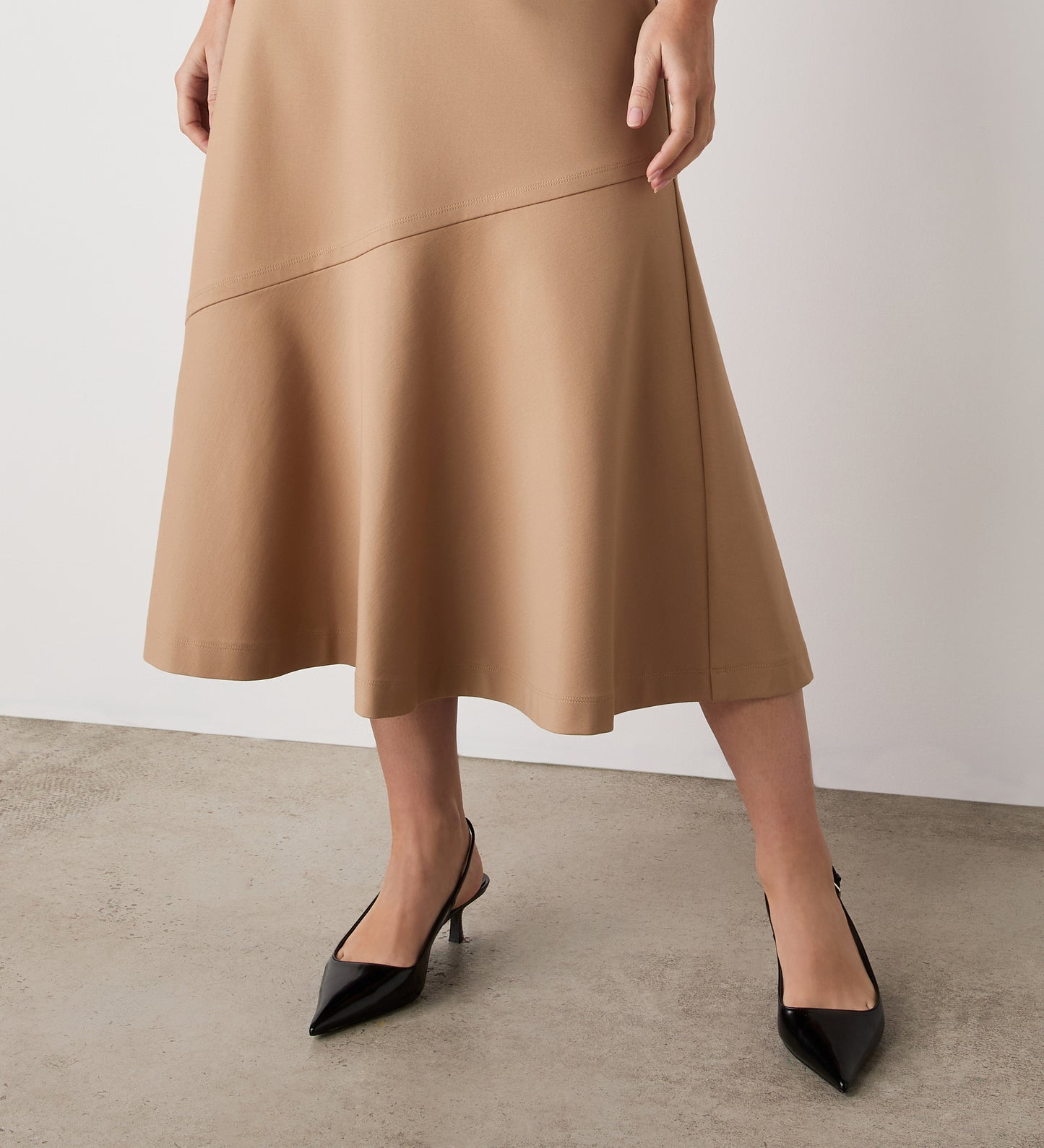 Model showcases the Grace Camel Ponte Jersey Midi Skirt—an effortlessly classic, tan, knee-length skirt—standing on a concrete floor against a plain white wall, with only the lower half of their body visible.