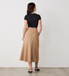 A woman faces a white wall, styled in a black short-sleeve top and the Grace Camel Ponte Jersey Midi Skirt, featuring high waist and asymmetrical seam. Black sandals and loose hair finish her timeless, versatile look.