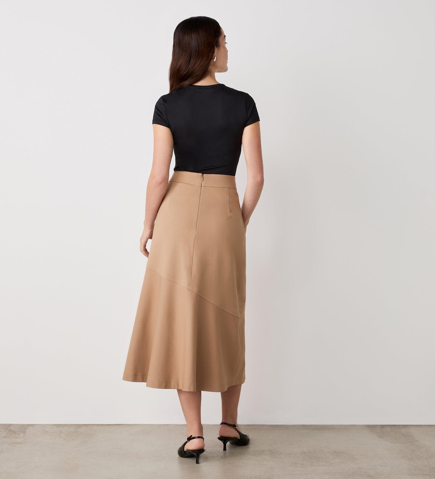 A woman faces a white wall, styled in a black short-sleeve top and the Grace Camel Ponte Jersey Midi Skirt, featuring high waist and asymmetrical seam. Black sandals and loose hair finish her timeless, versatile look.