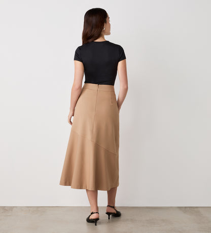 A woman faces a white wall, styled in a black short-sleeve top and the Grace Camel Ponte Jersey Midi Skirt, featuring high waist and asymmetrical seam. Black sandals and loose hair finish her timeless, versatile look.