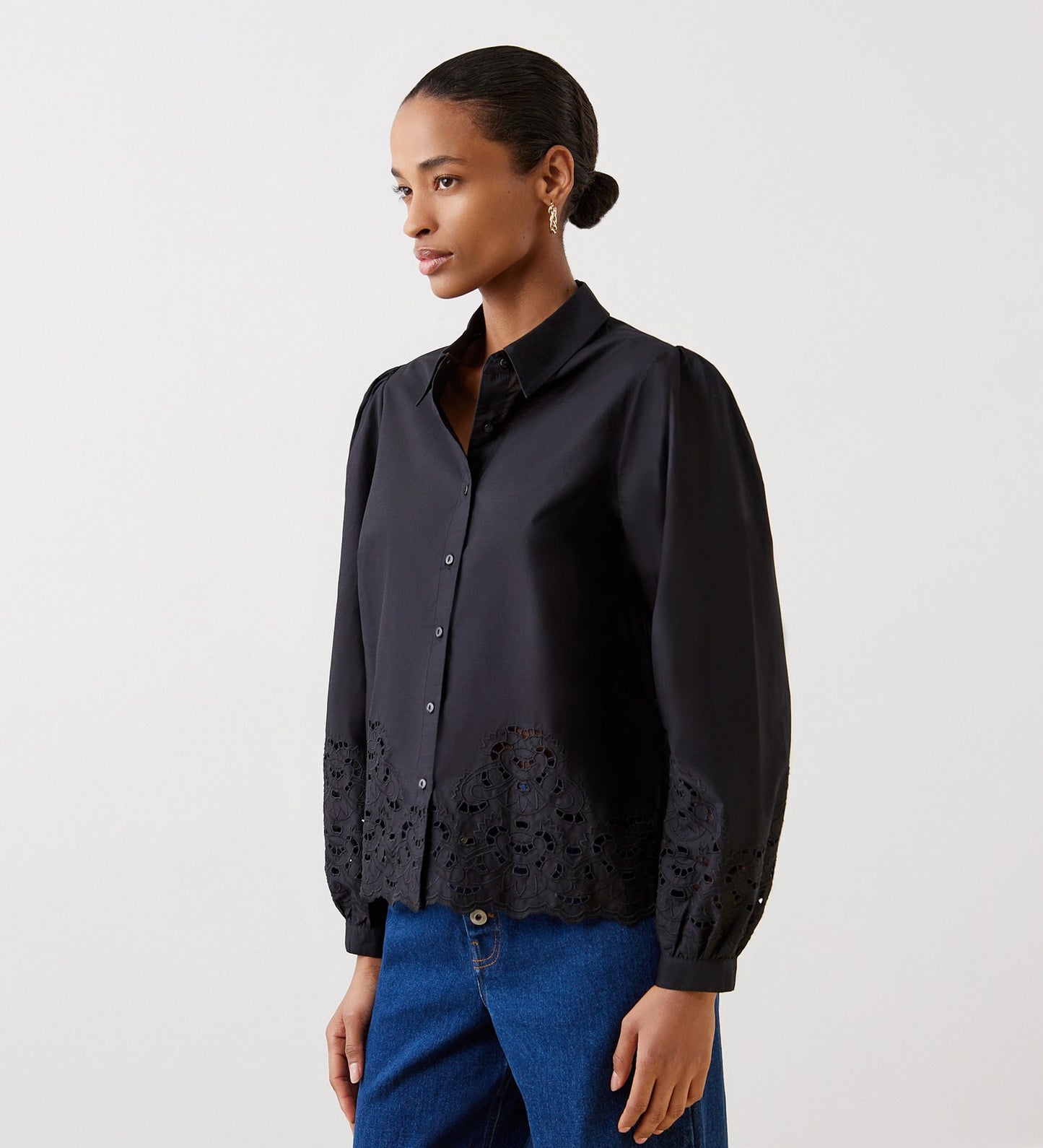 A woman wears the Jayne Black Broderie Cotton Shirt with blouson sleeves and embroidered hem and cuffs, paired with blue jeans, standing against a plain light background and looking to the side.
