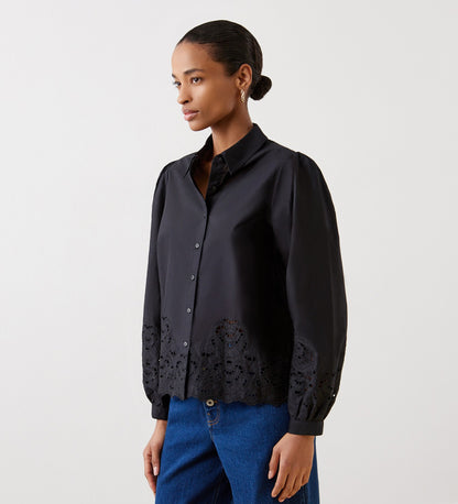 A woman wears the Jayne Black Broderie Cotton Shirt with blouson sleeves and embroidered hem and cuffs, paired with blue jeans, standing against a plain light background and looking to the side.