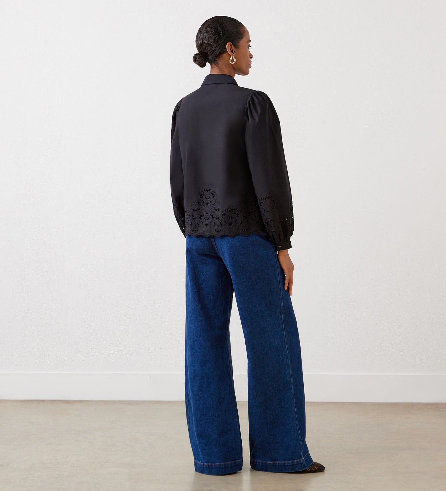 A woman with a low bun stands in a minimal, light room, wearing the Jayne Black Broderie Cotton Shirt with blouson sleeves and wide-leg blue jeans.