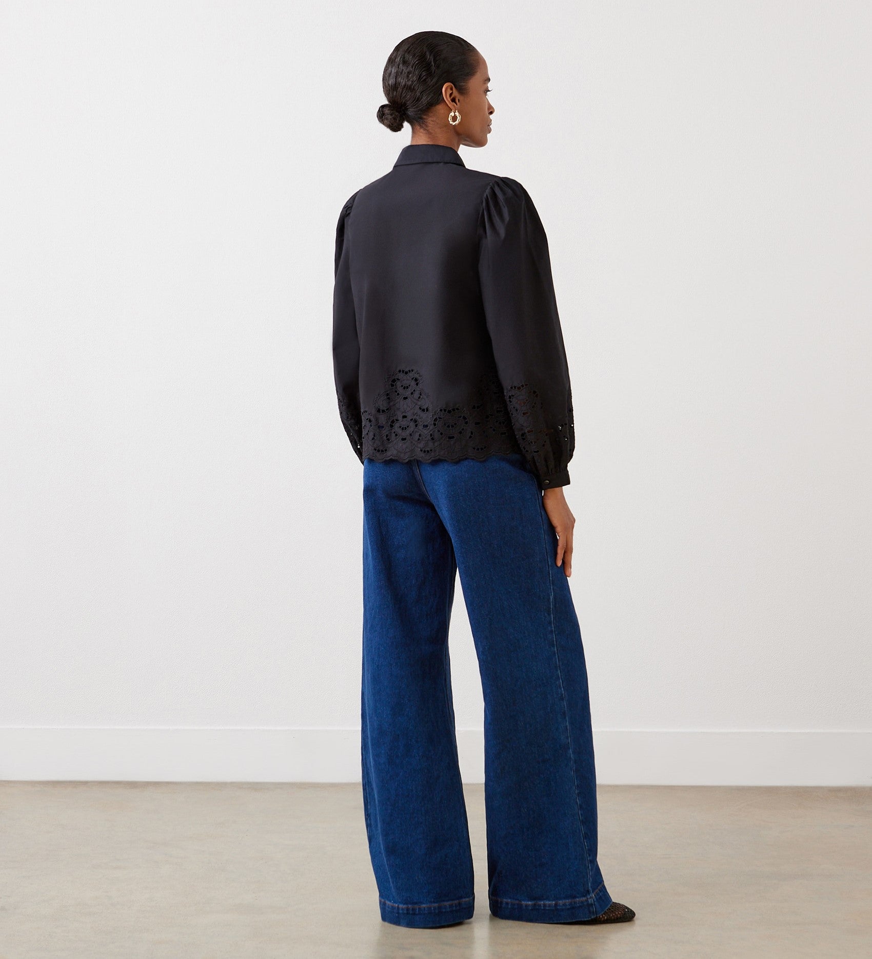 A woman with a low bun stands in a minimal, light room, wearing the Jayne Black Broderie Cotton Shirt with blouson sleeves and wide-leg blue jeans.