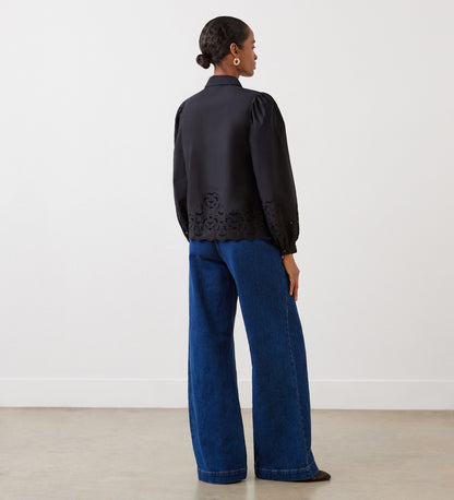 A woman with a low bun stands in a minimal, light room, wearing the Jayne Black Broderie Cotton Shirt with blouson sleeves and wide-leg blue jeans.