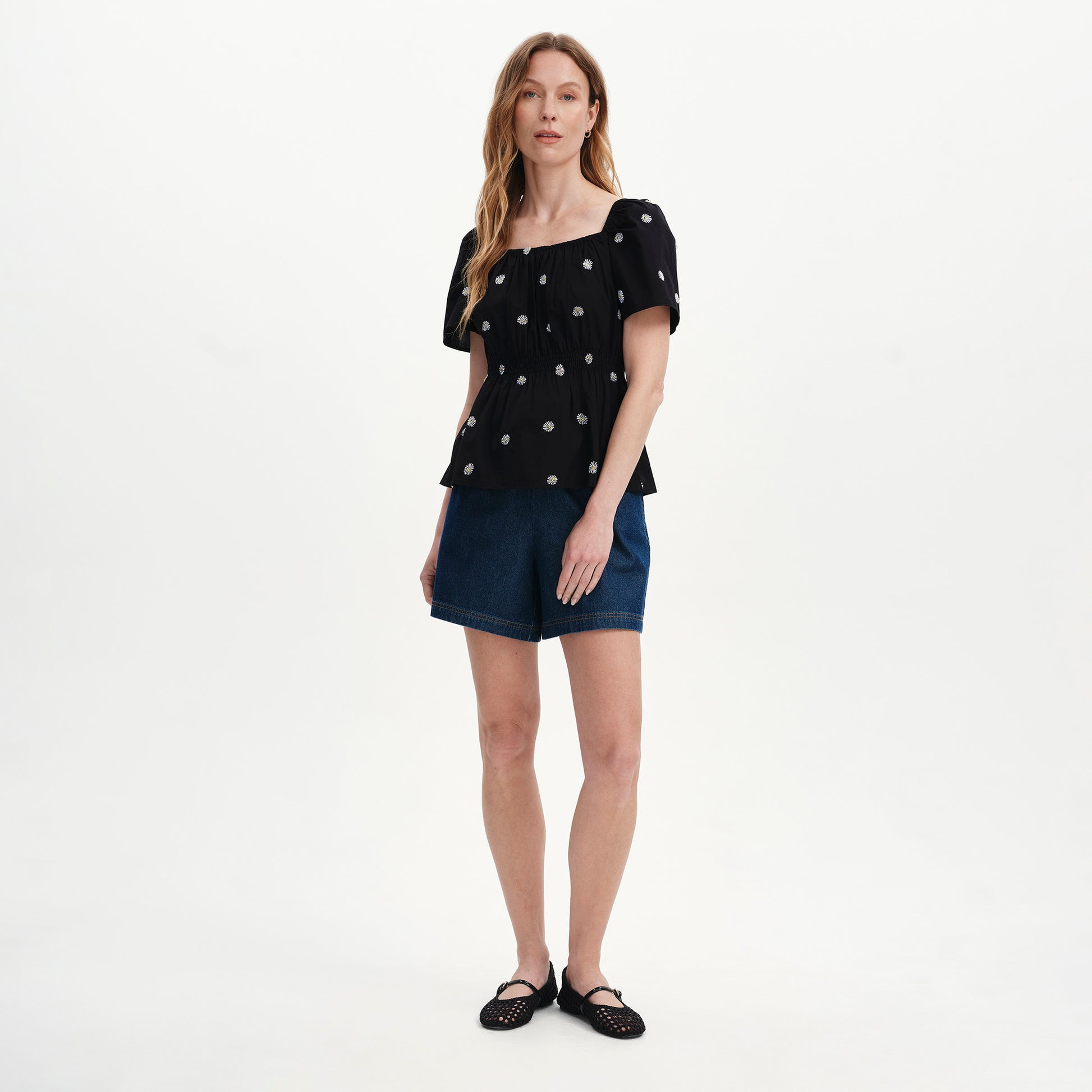 Woman wearing Holly Black Floral Embroidered Puff Sleeve Top and denim shorts on a white background