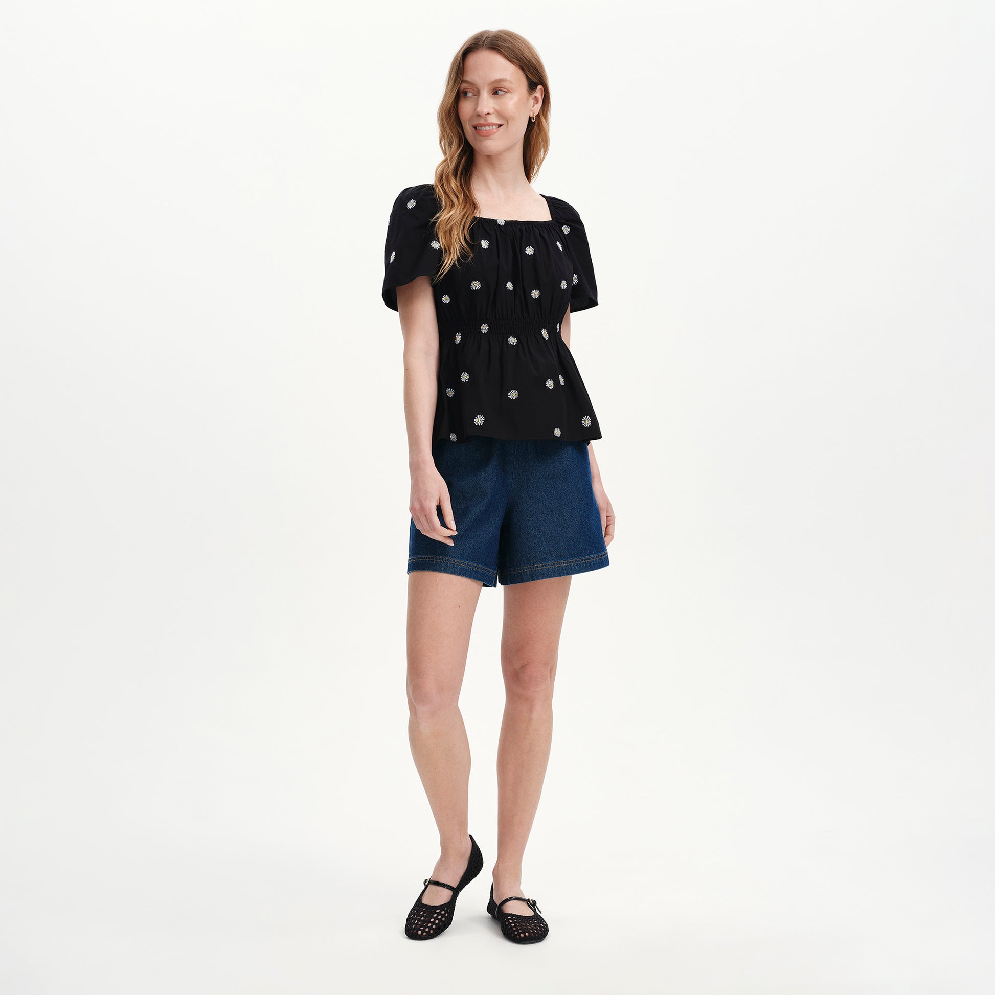Woman wearing Holly Black Floral Embroidered Puff Sleeve Top and denim shorts on a white background