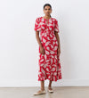 A woman stands indoors wearing the Isabella Red Paisley Tie Neck Crepe Midi Dress featuring an empire waist, short sleeves, and a ruffled hem. She accessorizes with hoop earrings, has her hair pulled back, and wears flat beige shoes.