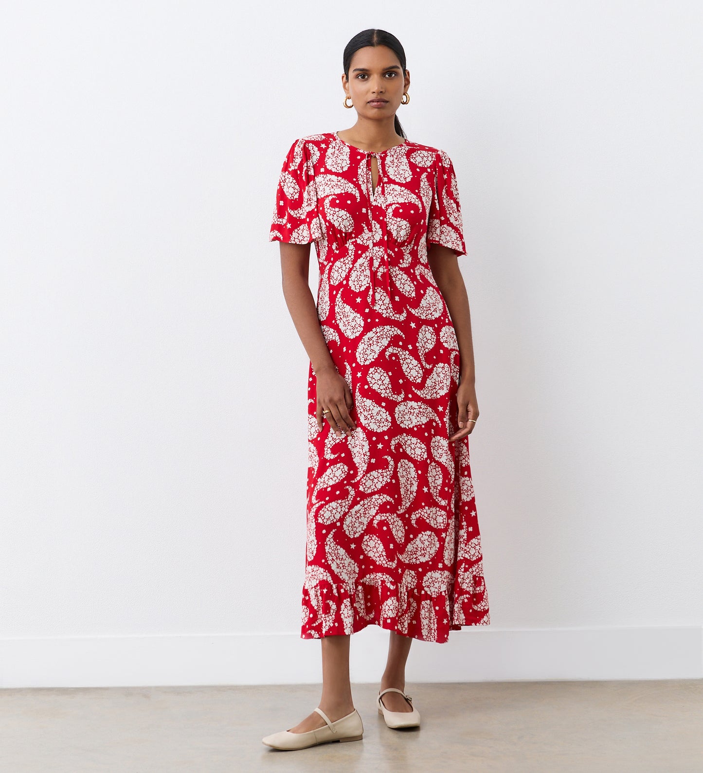 A woman stands indoors wearing the Isabella Red Paisley Tie Neck Crepe Midi Dress featuring an empire waist, short sleeves, and a ruffled hem. She accessorizes with hoop earrings, has her hair pulled back, and wears flat beige shoes.
