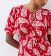 Wearing the Isabella Red Paisley Tie Neck Crepe Midi Dress, featuring an empire waist and tie neckline, a person stands against a white wall with their face partially out of frame.