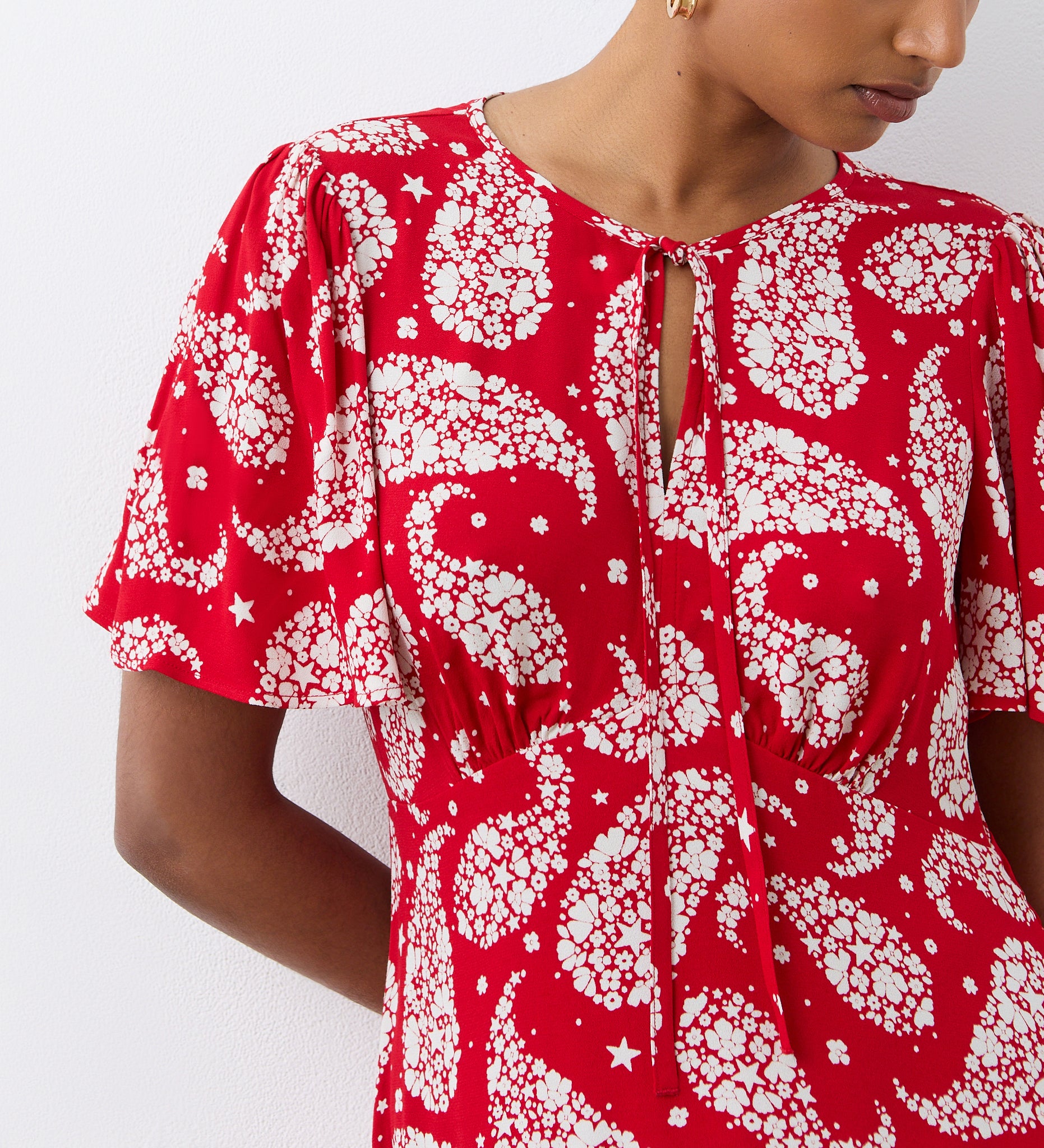 Wearing the Isabella Red Paisley Tie Neck Crepe Midi Dress, featuring an empire waist and tie neckline, a person stands against a white wall with their face partially out of frame.