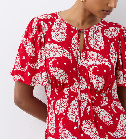 Wearing the Isabella Red Paisley Tie Neck Crepe Midi Dress, featuring an empire waist and tie neckline, a person stands against a white wall with their face partially out of frame.