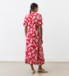 A woman with her dark hair tied back stands indoors facing away, wearing the Isabella Red Paisley Tie Neck Crepe Midi Dress and beige flats against a plain white wall.