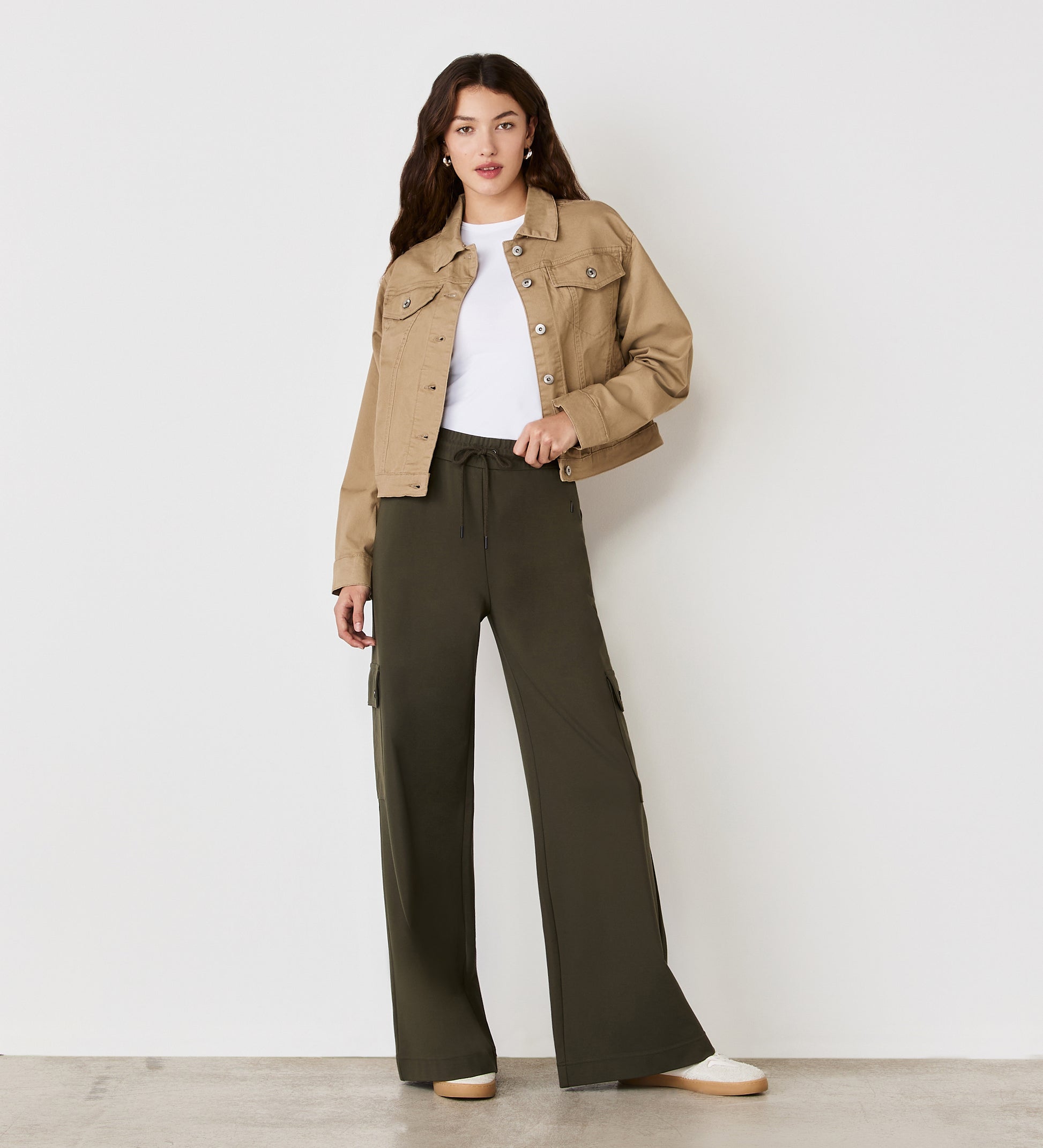 A woman with long, wavy brown hair stands against a white wall wearing the Isobel Mocha Denim Jacket over a white top, loose dark olive cargo pants, and white sneakers, looking toward the camera.