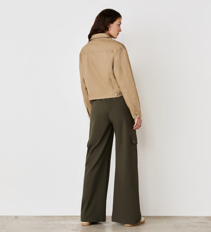 A woman stands facing away from the camera, wearing the Isobel Mocha Denim Jacket and olive green wide-leg pants with side pockets, against a plain white wall background.