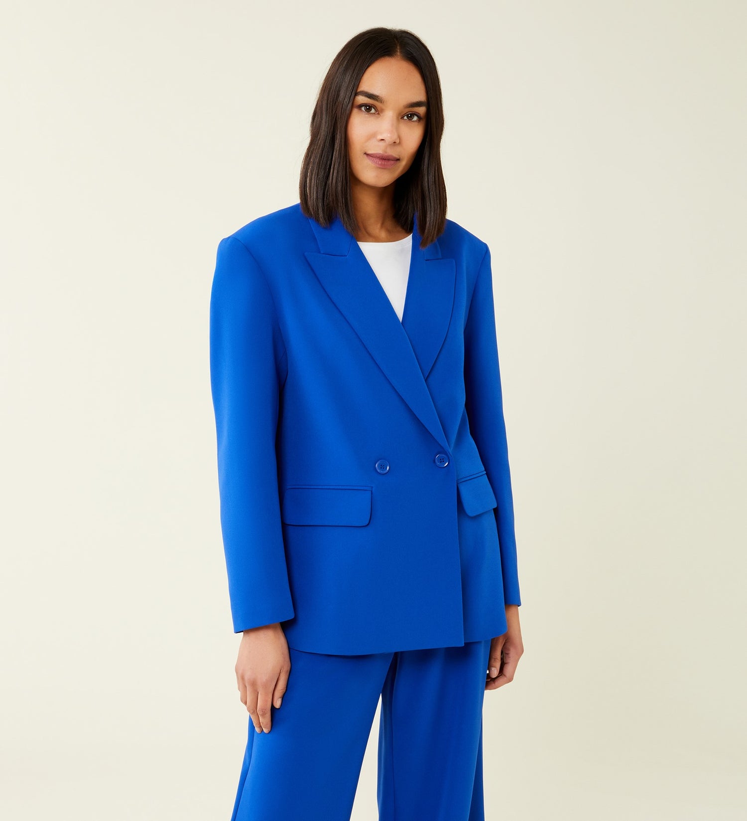 A woman with straight dark hair, wearing a Maeve Cobalt Blue Blazer and matching trousers over a white top, stands against a plain light background and looks at the camera with a slight smile.