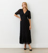 A woman with long blonde hair poses confidently against a white wall in the Loretta Black Lace Midi Dress, featuring short sleeves and a V-neckline, paired with black heels.