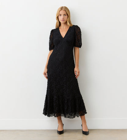 A woman with straight blonde hair stands against a white wall wearing the Loretta Black Lace Midi Dress, featuring a v-neckline, short puff sleeves, and flared hem. She completes her look with black pointed-toe heels.