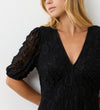 A woman with blonde hair wears the Loretta Black Lace Midi Dress, featuring short puffed sleeves and a deep v-neckline, while standing against a white textured wall. Her face is partially out of frame.