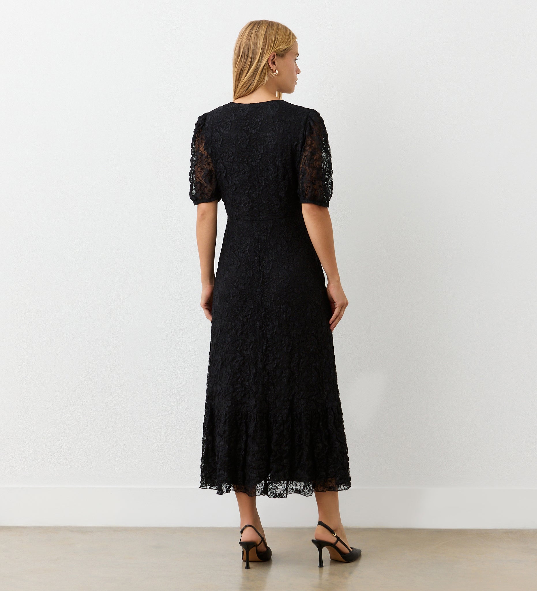 A woman faces away in a minimalist indoor space, wearing the Loretta Black Lace Midi Dress featuring a v-neckline and short puffed sleeves, paired with black heeled sandals against a light-colored wall and concrete floor.