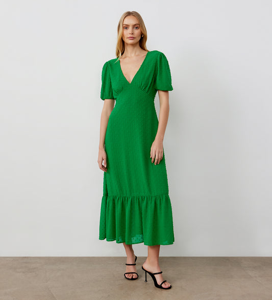 A woman stands against a plain white wall wearing the Loretta Green Georgette Midi Dress, featuring short puff sleeves, a deep V-neckline, crinkle texture, and ruffled hem. She completes her look with black heeled sandals.