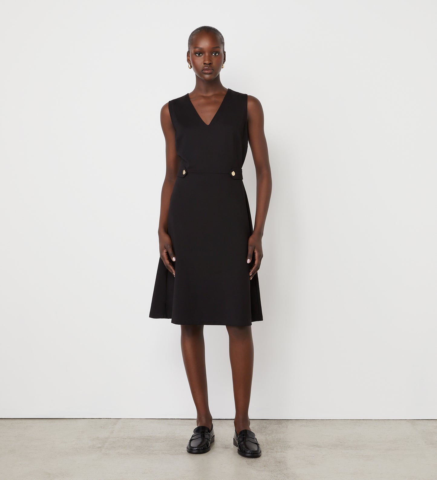 A woman stands against a plain white wall wearing the Lori Black Ponte Jersey Knee Length Dress, featuring a sleeveless v-neckline and two gold buttons at the waist, paired with black loafers.