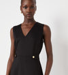 A woman with dark skin and gold hoop earrings wears the Lori Black Ponte Jersey Knee Length Dress—a sleeveless black dress with a v-neckline and gold buttons at the waist—standing against a plain white background.