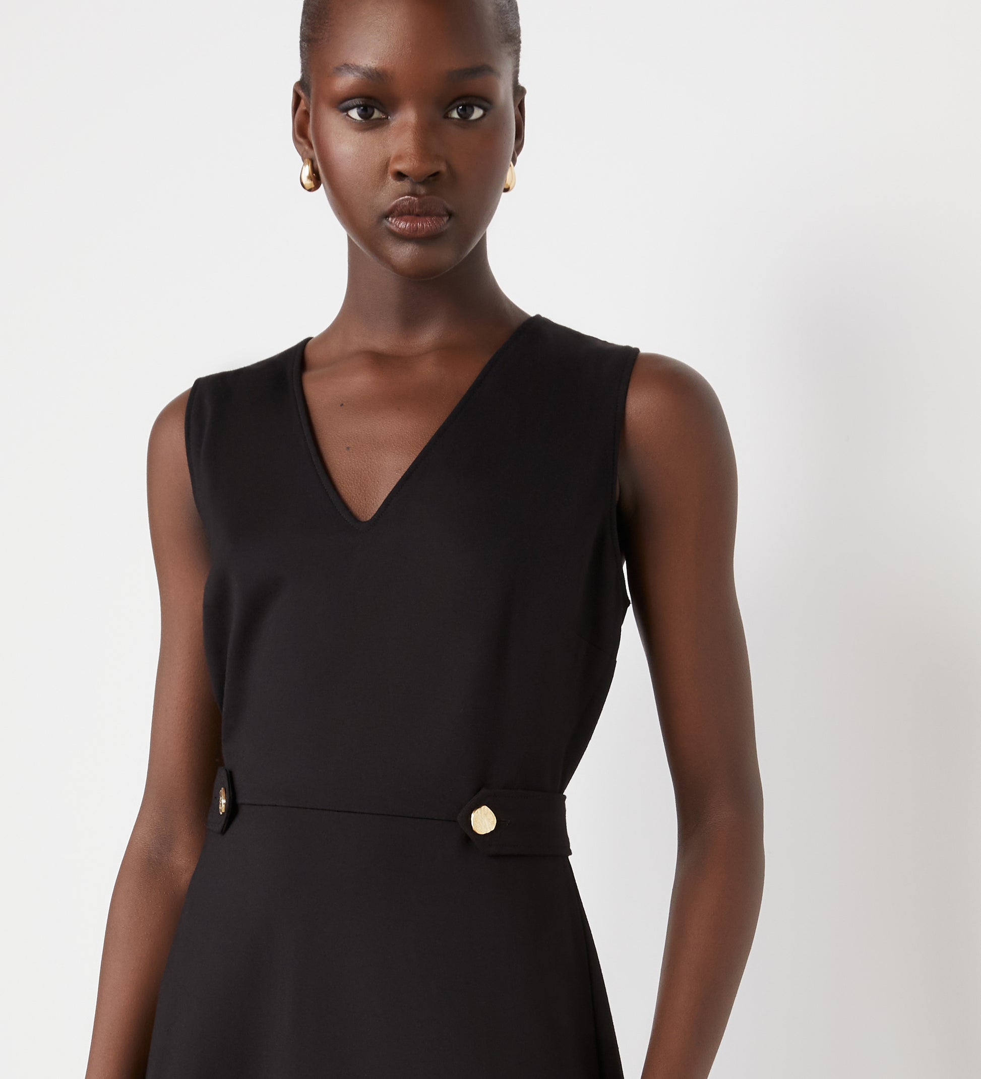 A woman with dark skin and gold hoop earrings wears the Lori Black Ponte Jersey Knee Length Dress—a sleeveless black dress with a v-neckline and gold buttons at the waist—standing against a plain white background.