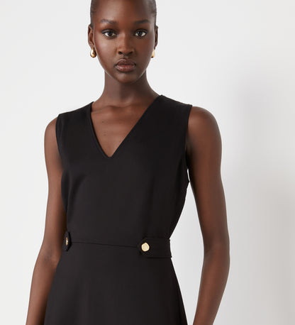 A woman with dark skin and gold hoop earrings wears the Lori Black Ponte Jersey Knee Length Dress—a sleeveless black dress with a v-neckline and gold buttons at the waist—standing against a plain white background.