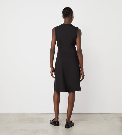 A person stands in a minimalist setting, facing away from the camera, wearing the Lori Black Ponte Jersey Knee Length Dress and black shoes against a white wall and concrete floor.