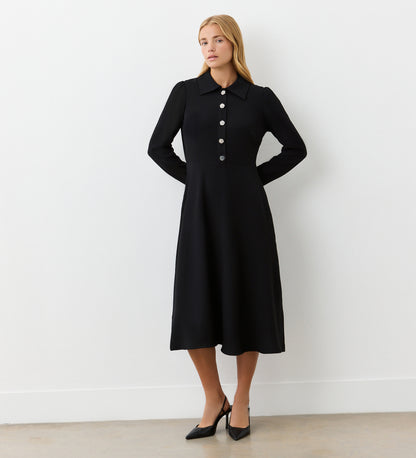 A woman stands against a white wall wearing the Lucia Black Midi Shirt Dress, featuring large buttons and a pointed collar—perfect for transitioning from office to after work. She finishes her look with black heels.