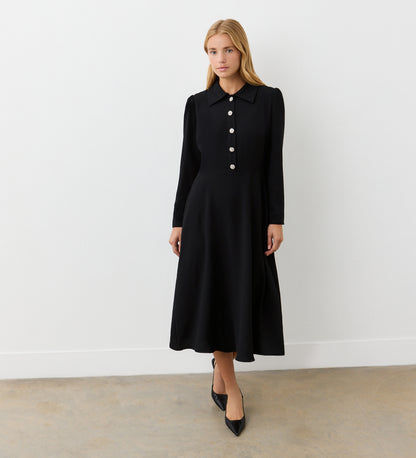 A woman with straight blonde hair stands against a white wall wearing the Lucia Black Midi Shirt Dress, featuring long sleeves, large front buttons, and a pointed collar. She completes her office-to-evening look with black pointed-toe shoes.
