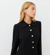 A woman with blonde hair models the Lucia Black Midi Shirt Dress, featuring pearl-look buttons and a pointed collar—ideal for office-to-evening style. She looks to the side against a plain white background.