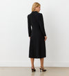 A woman with blonde hair faces away, wearing the Lucia Black Midi Shirt Dress and black heeled sandals—ideal for transitioning from office to after hours. The setting features a plain white wall and a light floor.