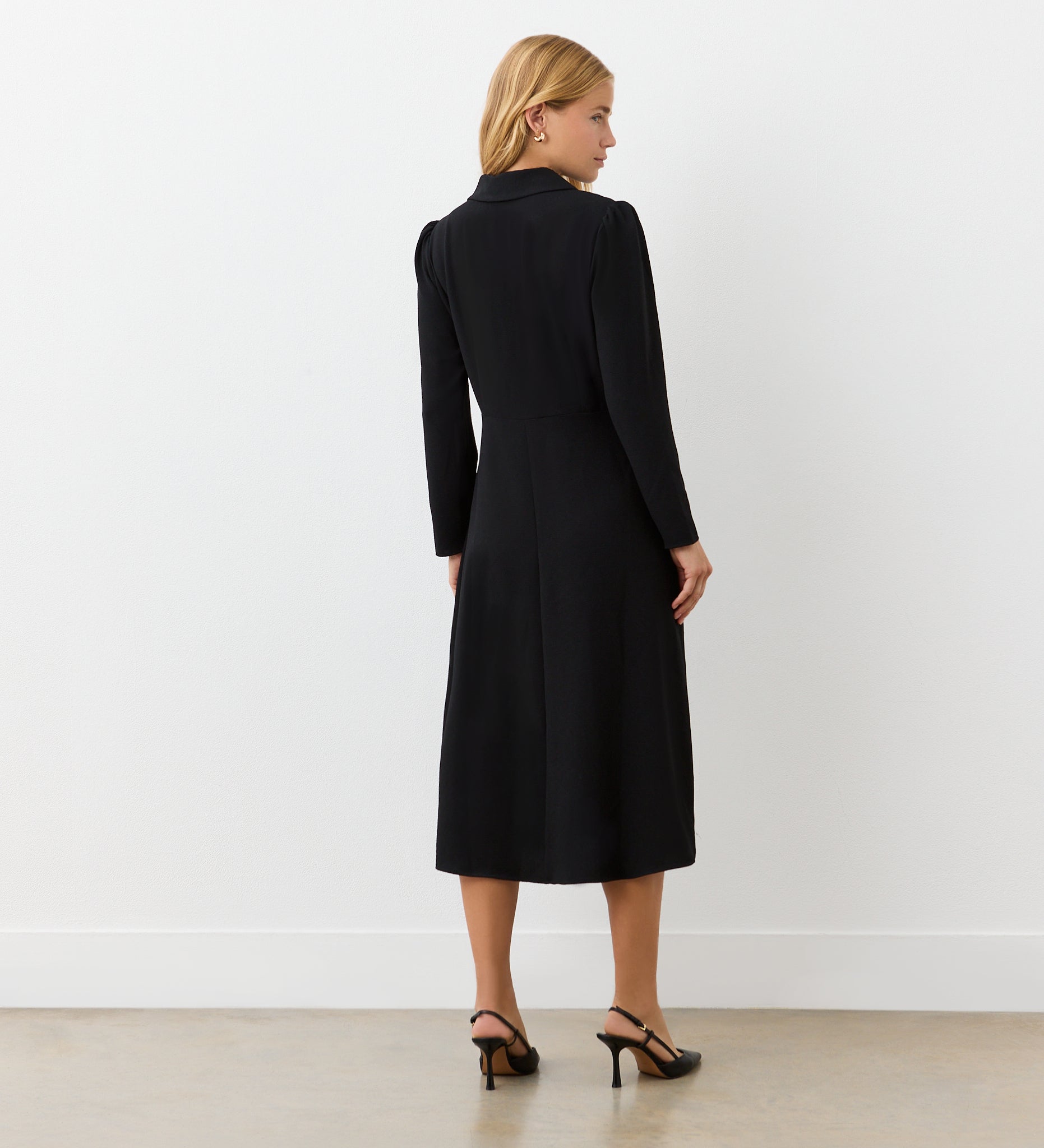A woman with blonde hair faces away, wearing the Lucia Black Midi Shirt Dress and black heeled sandals—ideal for transitioning from office to after hours. The setting features a plain white wall and a light floor.