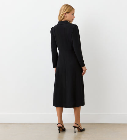 A woman with blonde hair faces away, wearing the Lucia Black Midi Shirt Dress and black heeled sandals—ideal for transitioning from office to after hours. The setting features a plain white wall and a light floor.