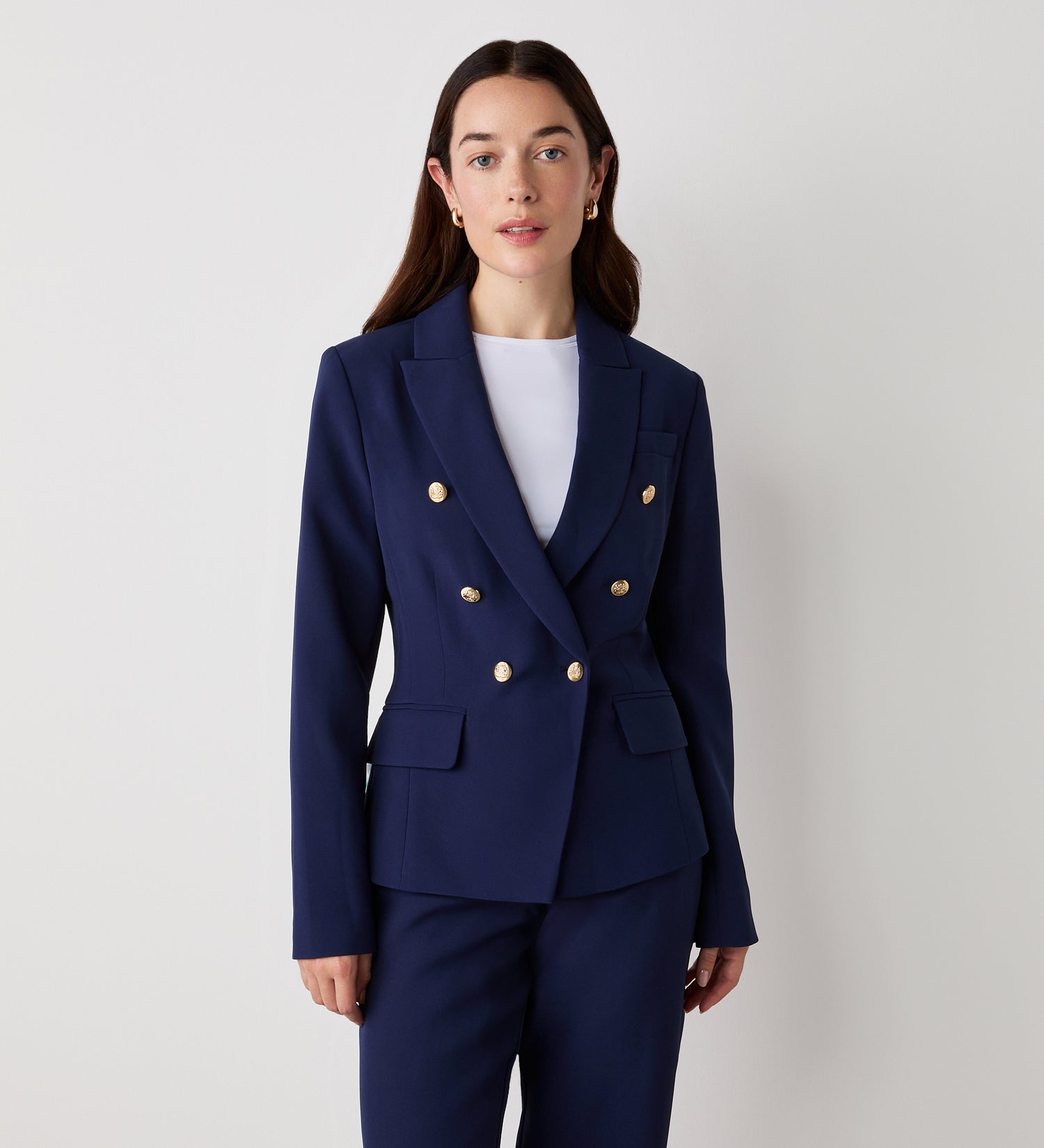 A woman with long brown hair and a neutral expression wears the Lydia Navy Blazer with gold button details and matching pants against a plain white background. She accessorizes with small earrings.