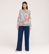 A woman with straight dark hair and a neutral expression wears the Lizzie Blue Floral Jersey Blouse, which features long sleeves and a frill stand collar, paired with wide-leg blue jeans against a plain light background.