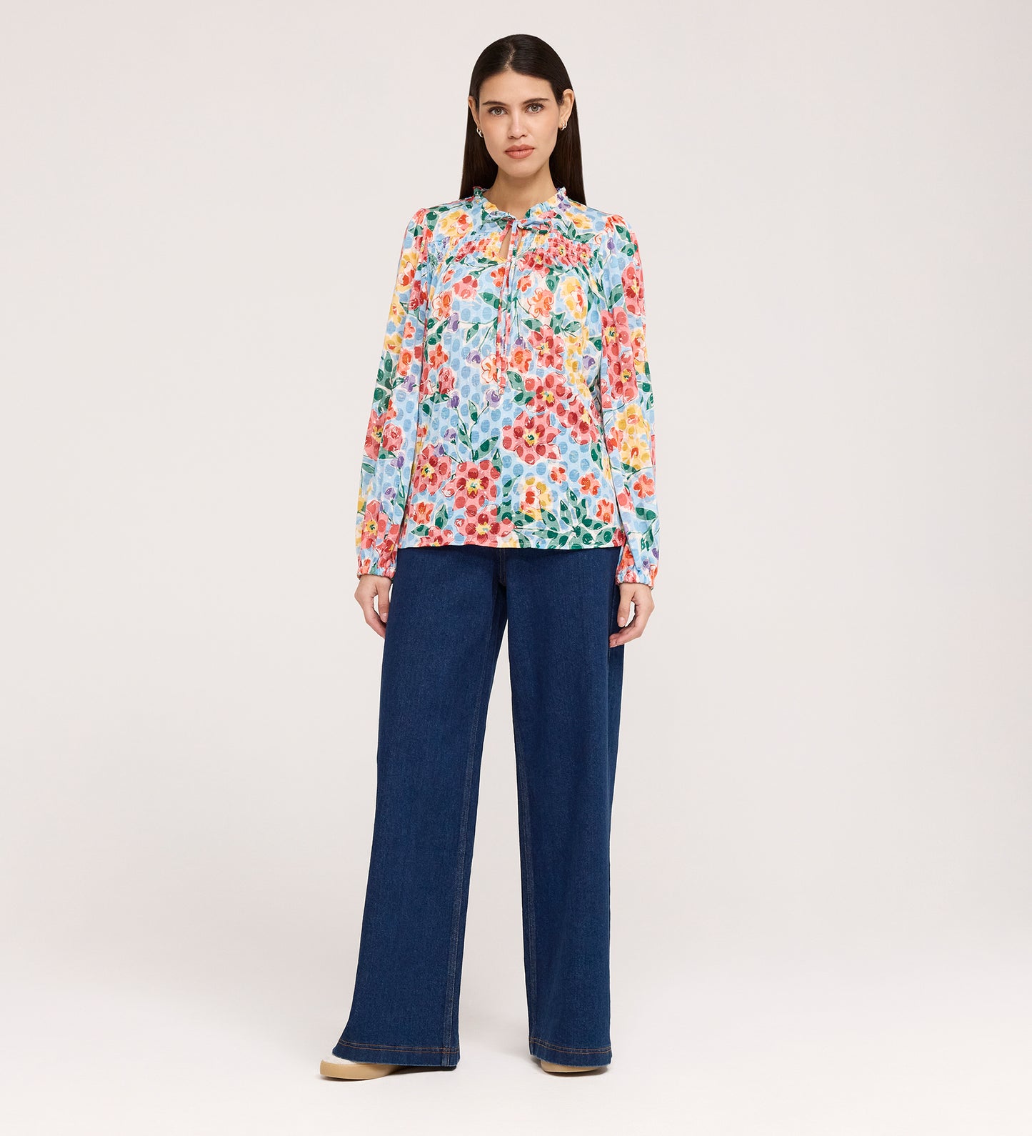 A woman with straight dark hair and a neutral expression wears the Lizzie Blue Floral Jersey Blouse, which features long sleeves and a frill stand collar, paired with wide-leg blue jeans against a plain light background.