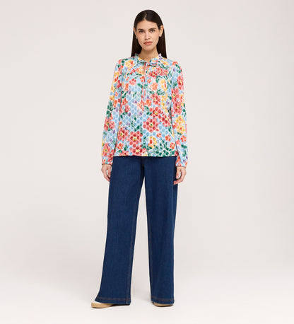A woman with straight dark hair and a neutral expression wears the Lizzie Blue Floral Jersey Blouse, which features long sleeves and a frill stand collar, paired with wide-leg blue jeans against a plain light background.