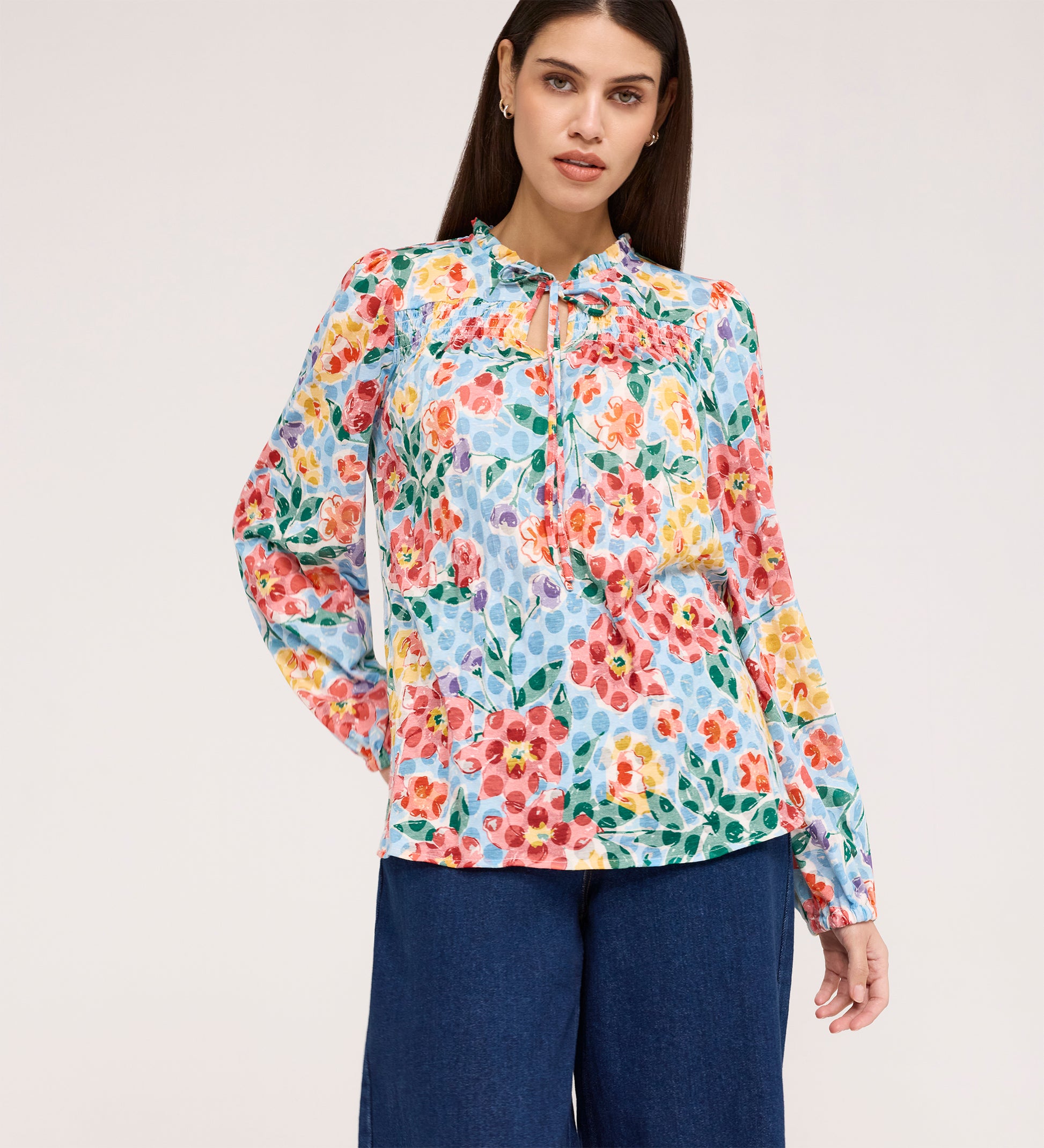 A woman stands against a plain light background wearing the Lizzie Blue Floral Jersey Blouse, featuring long sleeves, a tie neck, and a keyhole neckline, paired with blue wide-leg pants.