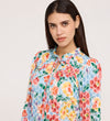 A woman with long dark hair wears the Lizzie Blue Floral Jersey Blouse, featuring a tie neck and frill stand collar, as she gazes neutrally at the camera against a plain light background.