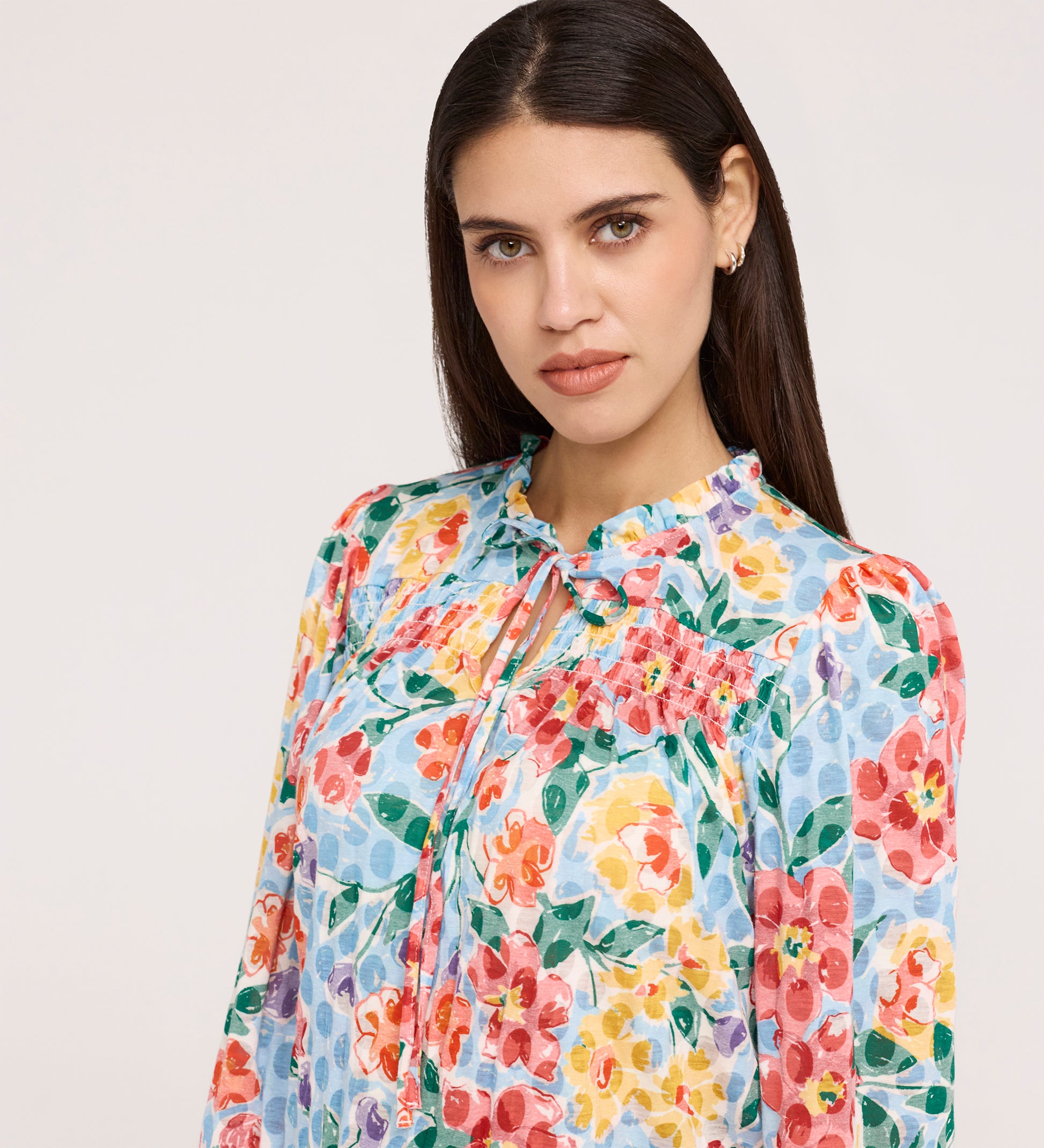 A woman with long dark hair wears the Lizzie Blue Floral Jersey Blouse, featuring a tie neck and frill stand collar, as she gazes neutrally at the camera against a plain light background.